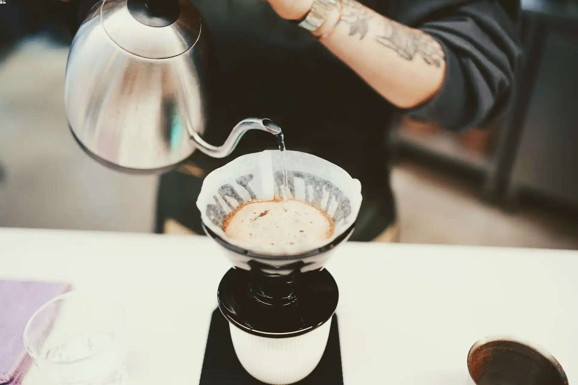 Tattooed arm pouring water from a kettle onto coffee grounds in a pour over filter set on a coffee mug on a scale.