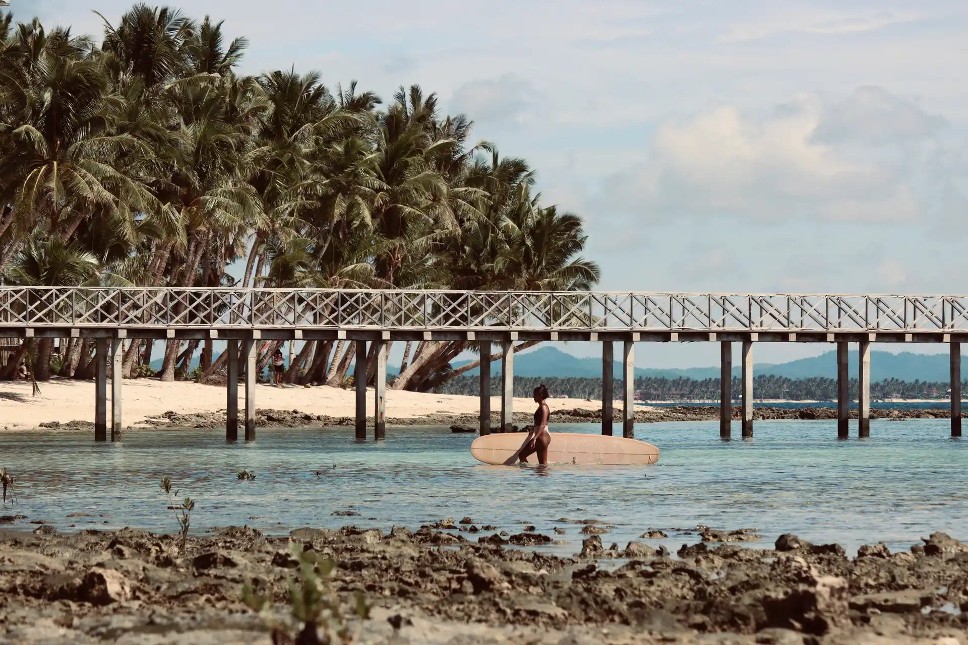 A woman standing in the water with a surfboard near a pier in the Philippines.