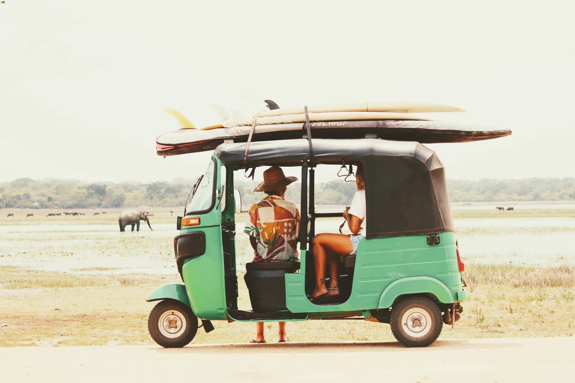 Two women in a green tuk tuk with surfboards strapped to the roof and an elephant in the background.