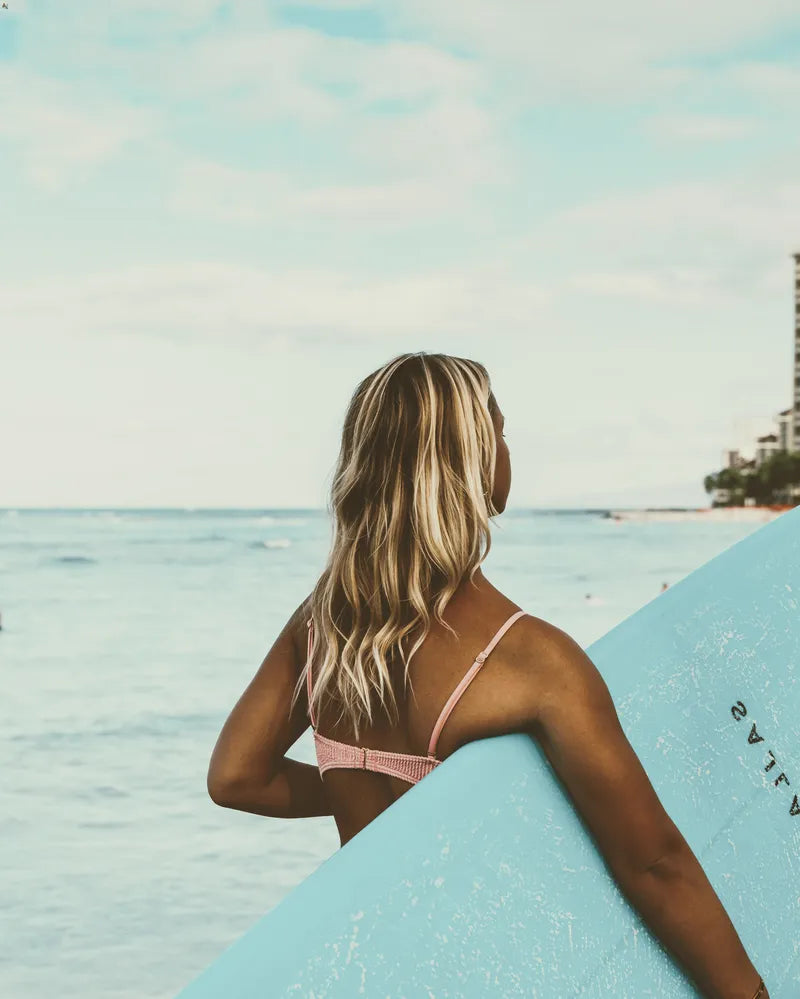 A blonde woman holding a blue surfboard on a sunny day at the ocean.