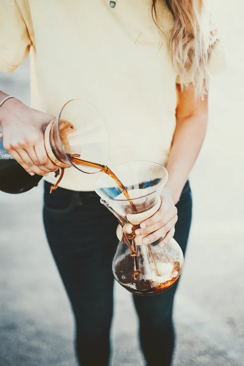 A woman wearing a pale yellow t-shirt pouring coffee from one Chemex to another outdoors.