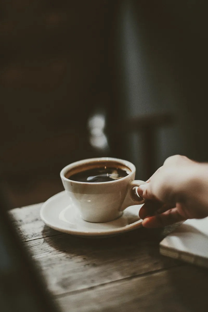 A hand grabbing a cup of dark espresso next to a journal in a moody setting.