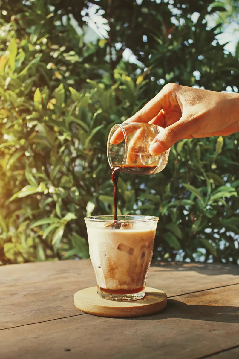 A hand pouring espresso into a glass filled with ice and milk on an outdoors table in the sun.