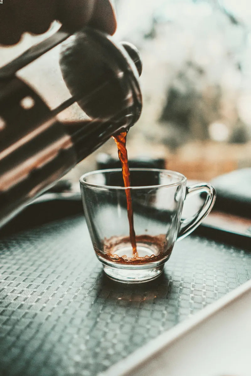 Coffee pouring from a French press into a glass mug on a green textured tray outdoors.