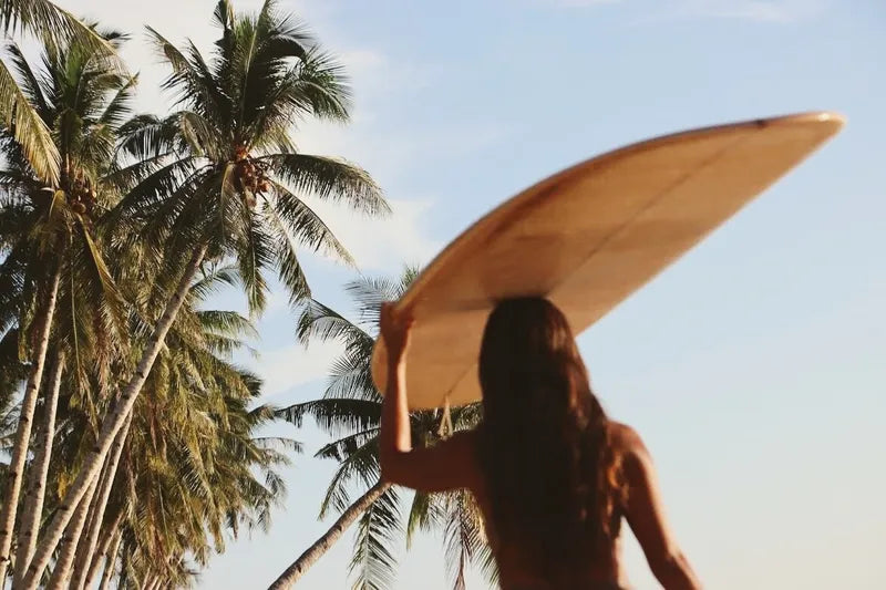 A woman balancing a surfboard on her head with one hand in the sun near palm trees.