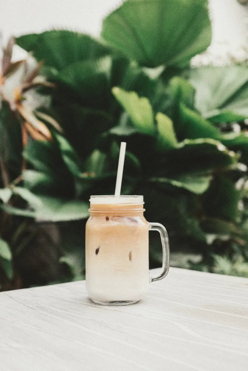 A mason jar mug of iced coffee with a straw on an table outdoors in the sun with plants in the background.