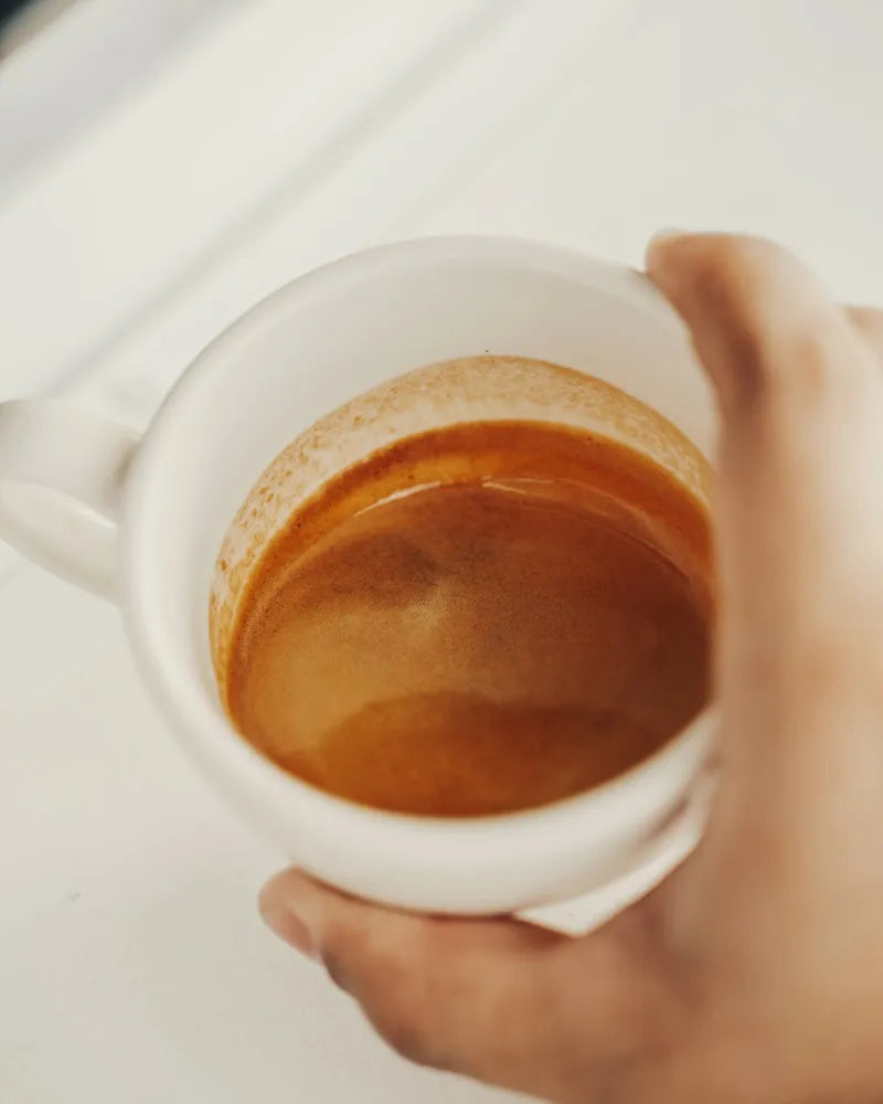 A hand holding a white mug of light espresso with frothy crema.