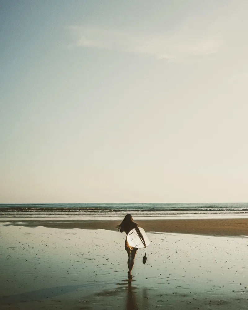 A woman carrying a surfboard into the ocean at sunset.