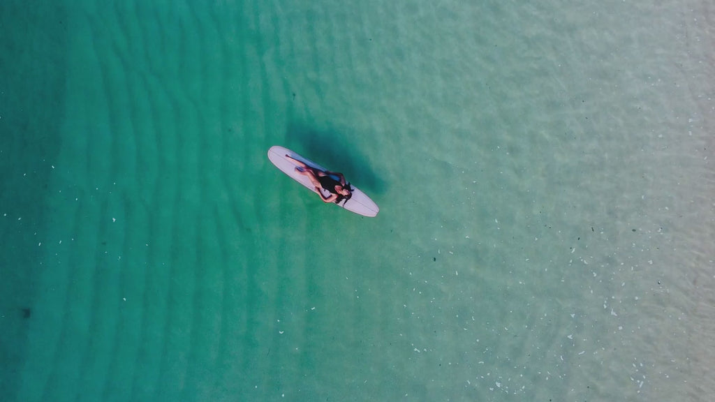 Drone footage of a woman lying on a surfboard in the calm shallow blue ocean near the beach.